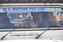 A man inside a vehicle with his hands pressed against the windshield. He appears to be making a playful or surprised expression. The vehicle has a sign with the name 'K. S. Motors Pvt. Ltd.' on the windshield and is a Mahindra model. Another person is seated next to the man inside the vehicle.