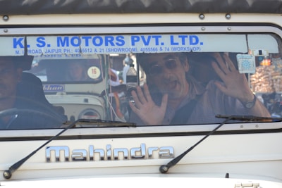 A man inside a vehicle with his hands pressed against the windshield. He appears to be making a playful or surprised expression. The vehicle has a sign with the name 'K. S. Motors Pvt. Ltd.' on the windshield and is a Mahindra model. Another person is seated next to the man inside the vehicle.