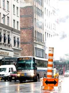 A busy urban street with a public bus in the center. The surrounding tall buildings exhibit a mix of modern and older architecture with brick and concrete facades. A prominent orange and white construction barrier with arrows sits in the foreground, indicating traffic direction. Steam rises from a vent, creating a slightly hazy atmosphere. Several other vehicles, including a taxi and a van, are visible on the wet roadway.