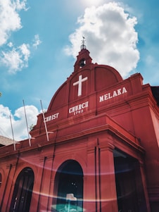 A vibrant red building with architectural features typical of colonial-era churches stands against a bright blue sky. The facade displays the words 'Christ Church Melaka 1753' in white, along with a prominent white cross at the top. The sky is dotted with fluffy white clouds, and the sunlight creates a lens flare effect at the bottom of the image.