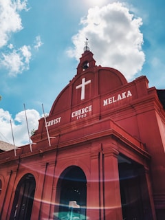 A vibrant red building with architectural features typical of colonial-era churches stands against a bright blue sky. The facade displays the words 'Christ Church Melaka 1753' in white, along with a prominent white cross at the top. The sky is dotted with fluffy white clouds, and the sunlight creates a lens flare effect at the bottom of the image.
