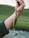 A close-up of a hand holding a fishing line with a small fish attached. The fish has a speckled pattern with brown spots and a mix of golden and silver scales. The background features a blurred landscape of green grass and a flowing stream.