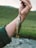A close-up of a hand holding a fishing line with a small fish attached. The fish has a speckled pattern with brown spots and a mix of golden and silver scales. The background features a blurred landscape of green grass and a flowing stream.