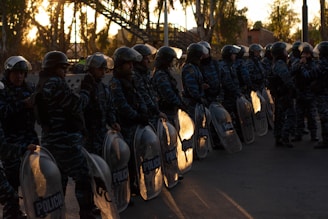 A group of uniformed riot police officers standing in a line, each holding a clear shield. They are wearing helmets and camouflage uniforms, with a backdrop of trees and structures visible against a setting sun.