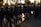 A group of uniformed riot police officers standing in a line, each holding a clear shield. They are wearing helmets and camouflage uniforms, with a backdrop of trees and structures visible against a setting sun.