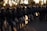 A group of uniformed riot police officers standing in a line, each holding a clear shield. They are wearing helmets and camouflage uniforms, with a backdrop of trees and structures visible against a setting sun.