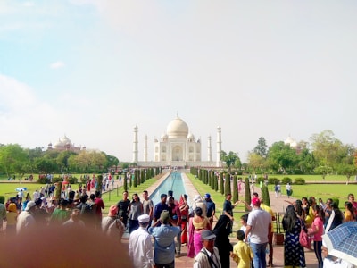 A large crowd of people gathers near the famous white marble mausoleum set against a clear blue sky. The symmetrical structure features a central dome and four minarets, surrounded by lush green gardens and a long reflecting pool. Tourists of various ages and backgrounds are taking photos and walking along the garden path.