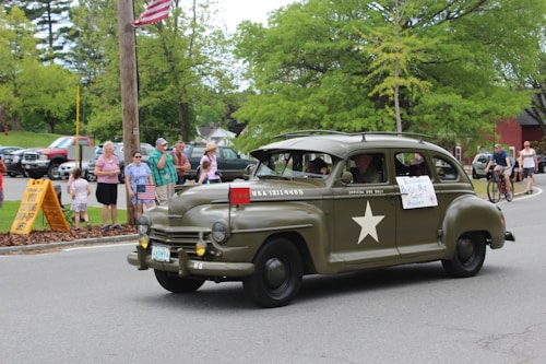 An olive green vintage army vehicle with a white star on the side drives down a street. People, including adults and children, are gathered along the sidewalk, some holding American flags. The background features lush green trees and parked cars. A sign reading 'Distinguished Boston Rot. Guest Holder' is taped to the car window.