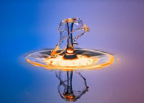 A dynamic splash of water creates a symmetrical and intricate pattern as it rises and falls, with reflections visible on the surface. The colors transition smoothly from deep blue on the left to a warm, glowing orange on the right, highlighting the fluid motion and delicate structure of the splash.