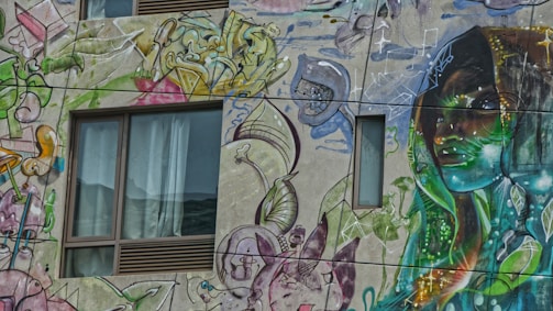 A vibrant mural covers the side of a building, featuring abstract designs and a detailed portrait. The artwork incorporates various colors and shapes, including organic forms and geometric patterns, surrounding a human-like figure. The windows on the building are seamlessly integrated into the mural, adding depth to the scene.