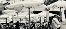 An outdoor cafe setting with numerous people seated under large umbrellas. The umbrellas display branding, and people appear to be engaged in conversation, enjoying a relaxed atmosphere. The scene is in black and white, adding a classic and timeless feel.
