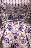 A detailed close-up of an intricately embroidered garment, featuring floral patterns adorned with silver and blue beadwork. The fabric is satin-like, with a lustrous sheen giving it an opulent appearance. The embroidery is rich in detail, combining swirling designs with symmetrical flower motifs.