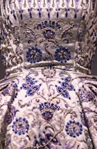 A detailed close-up of an intricately embroidered garment, featuring floral patterns adorned with silver and blue beadwork. The fabric is satin-like, with a lustrous sheen giving it an opulent appearance. The embroidery is rich in detail, combining swirling designs with symmetrical flower motifs.