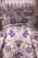 A detailed close-up of an intricately embroidered garment, featuring floral patterns adorned with silver and blue beadwork. The fabric is satin-like, with a lustrous sheen giving it an opulent appearance. The embroidery is rich in detail, combining swirling designs with symmetrical flower motifs.