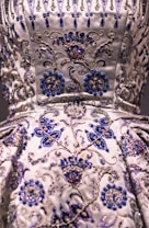 A detailed close-up of an intricately embroidered garment, featuring floral patterns adorned with silver and blue beadwork. The fabric is satin-like, with a lustrous sheen giving it an opulent appearance. The embroidery is rich in detail, combining swirling designs with symmetrical flower motifs.