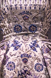 A detailed close-up of an intricately embroidered garment, featuring floral patterns adorned with silver and blue beadwork. The fabric is satin-like, with a lustrous sheen giving it an opulent appearance. The embroidery is rich in detail, combining swirling designs with symmetrical flower motifs.