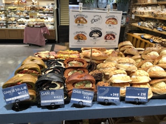 A display of various types of bread in a bakery, including dark and light loaves with different fillings. Signs with prices and names in Japanese are placed in front of the bread. Shelves filled with more baked goods are visible in the background.