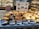 A display of various types of bread in a bakery, including dark and light loaves with different fillings. Signs with prices and names in Japanese are placed in front of the bread. Shelves filled with more baked goods are visible in the background.
