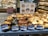 A display of various types of bread in a bakery, including dark and light loaves with different fillings. Signs with prices and names in Japanese are placed in front of the bread. Shelves filled with more baked goods are visible in the background.