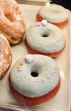 A collection of donuts on display, featuring three donuts with light blue frosting and decorative toppings such as small white hearts and silver pearls. There is also a classic donut adorned with powdered sugar to the side.