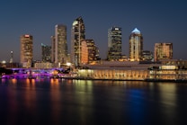 A city skyline at dusk featuring several tall buildings illuminated with lights. The Tampa Convention Center is prominently positioned in the middle ground, and a body of water in the foreground reflects the colorful lights of the cityscape. The sky is a deep blue as night falls.
