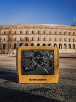 An old, yellow CRT television with a shattered screen is placed on an empty paved surface. Behind it, a large, round, historical building with arches is visible against a clear blue sky.