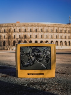 An old, yellow CRT television with a shattered screen is placed on an empty paved surface. Behind it, a large, round, historical building with arches is visible against a clear blue sky.