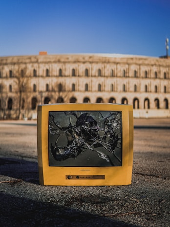 An old, yellow CRT television with a shattered screen is placed on an empty paved surface. Behind it, a large, round, historical building with arches is visible against a clear blue sky.