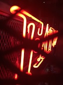 A brightly illuminated red neon sign with stylized script, placed against a mesh or grid background. The neon tubes are prominently glowing and seem partially obscured by overlapping shadow patterns.