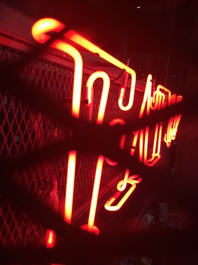 A brightly illuminated red neon sign with stylized script, placed against a mesh or grid background. The neon tubes are prominently glowing and seem partially obscured by overlapping shadow patterns.