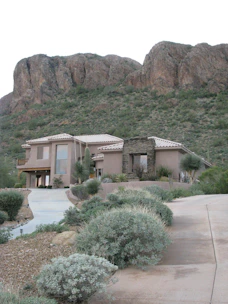 A modern house with a stucco exterior is situated at the base of a rocky hillside surrounded by desert vegetation. The landscape includes various desert plants and shrubs along a concrete driveway leading to the entrance of the house. The background features large rock formations characteristic of a desert environment.