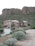 A modern house with a stucco exterior is situated at the base of a rocky hillside surrounded by desert vegetation. The landscape includes various desert plants and shrubs along a concrete driveway leading to the entrance of the house. The background features large rock formations characteristic of a desert environment.