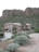 A modern house with a stucco exterior is situated at the base of a rocky hillside surrounded by desert vegetation. The landscape includes various desert plants and shrubs along a concrete driveway leading to the entrance of the house. The background features large rock formations characteristic of a desert environment.