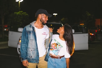 A man and woman stand closely together in a parking lot at night, looking at each other playfully. The man is wearing a denim jacket, a white t-shirt, and a baseball cap, while the woman is wearing a colorful t-shirt and denim shorts. Behind them, a parked car has its doors open, and the scene is illuminated by streetlights.