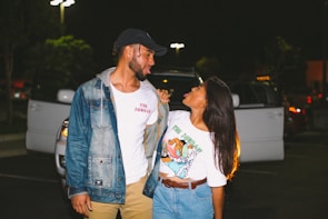 A man and woman stand closely together in a parking lot at night, looking at each other playfully. The man is wearing a denim jacket, a white t-shirt, and a baseball cap, while the woman is wearing a colorful t-shirt and denim shorts. Behind them, a parked car has its doors open, and the scene is illuminated by streetlights.
