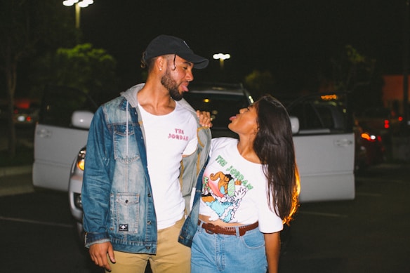 A man and woman stand closely together in a parking lot at night, looking at each other playfully. The man is wearing a denim jacket, a white t-shirt, and a baseball cap, while the woman is wearing a colorful t-shirt and denim shorts. Behind them, a parked car has its doors open, and the scene is illuminated by streetlights.