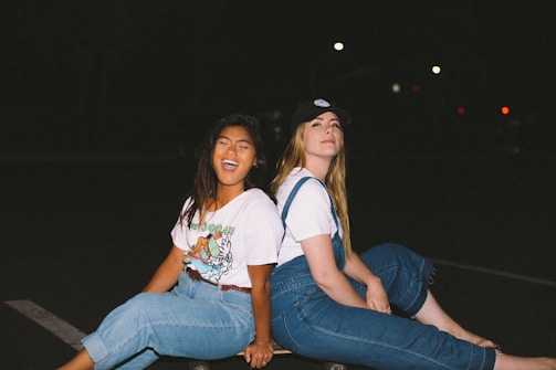 Two young women sit back-to-back on a skateboard in an outdoor setting at night. Both are wearing casual outfits consisting of jeans and white tops. One woman has long dark hair and is smiling widely, appearing joyful and carefree. The other woman has long lighter hair, wears a cap, and has a relaxed expression.