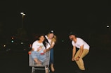 Three young adults are having fun in a parking lot at night. One person is sitting in a shopping cart while another pushes it and a third person holds onto the cart laughing. There are streetlights in the background, providing light to the scene.