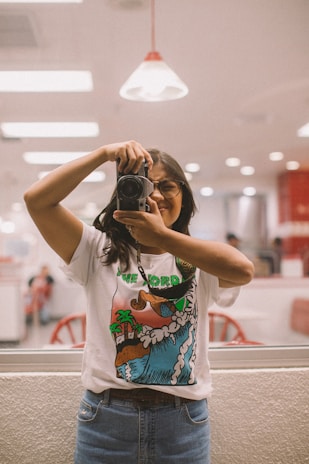 A person with long dark hair and glasses is holding a camera, taking a photo in what appears to be a brightly lit indoor setting. They are wearing a graphic t-shirt with a colorful design featuring nature elements like trees and waves, and a pair of blue jeans. Behind them, there are reflections of red chairs and tables.