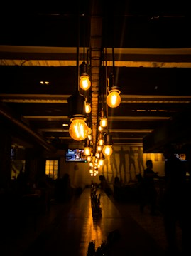 A dimly lit interior scene featuring a row of vintage hanging lightbulbs casting a warm amber glow. The room is adorned with dark furniture and a long table leading towards a group of people in the background. The walls reflect the light, creating a cozy atmosphere.