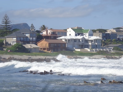 Several houses are perched on a grassy hill overlooking a rocky coastline. The ocean waves crash against the shore, creating white foam. The architecture of the houses varies, with a mix of modern and traditional styles. In the background, tree-covered hills are visible under a partly cloudy sky.