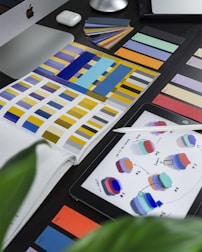 A workspace featuring a variety of color swatches and design tools. There is a desktop computer and open notebooks displaying different color palettes. Nearby, an iPad with a stylus shows an application with color-related graphics.