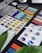A workspace featuring a variety of color swatches and design tools. There is a desktop computer and open notebooks displaying different color palettes. Nearby, an iPad with a stylus shows an application with color-related graphics.