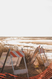 Traffic barriers with red and white stripes are set up near a sandy area with overgrown grass. The background features a bright, reflective body of water under a clear sky, suggesting an open and potentially desolate setting.