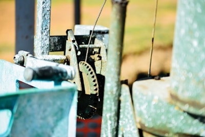 A close-up view of mechanical components, including a gear and a metal rod. The elements display signs of wear and rust, suggesting exposure to the elements. The background is blurred and features warm colors, possibly indicating an outdoor environment.