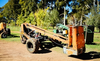 A rusty orange farm machine sits on a dirt path surrounded by green trees and grass. The machinery appears old and weathered, with visible wear on its metal surfaces. Behind the main machine, other pieces of farming equipment are partially visible among the foliage.