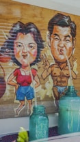 A humorous illustration depicts two cartoonish characters with exaggerated features. The female character wears a red top and blue shorts, while the male character is muscular and wearing a minimal swimsuit. Both characters are connected to green glass bottles via long straws.