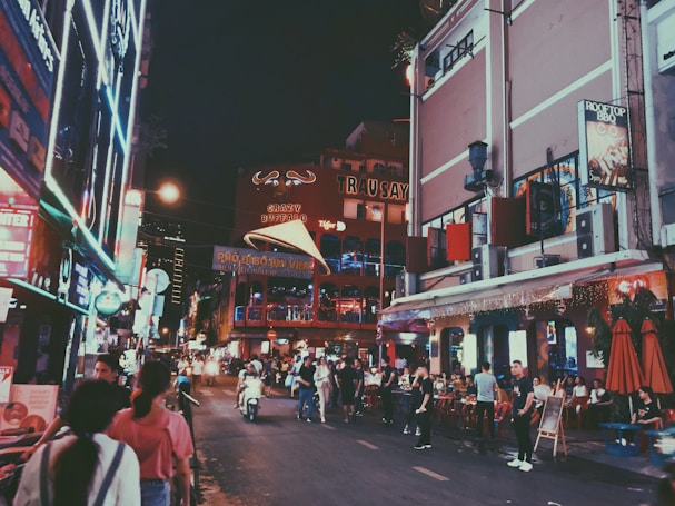 A lively city street bustling with people walking and sitting outside restaurants. Neon lights illuminate the scene, with several signs and advertisements visible on the buildings. The street has a vibrant and busy atmosphere with a mix of locals and tourists.
