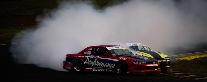 Two cars are engaged in a high-speed drift around a track, producing a large cloud of smoke. The car in the foreground is red with visible sponsorship decals, while the one in the background is yellow and black. The scene is intense and dynamic, capturing the excitement of motorsport.