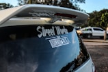 A rear view of a vehicle featuring a large spoiler and a decal on the back window with the text 'Street Works'. In the background, another vehicle, possibly a pickup truck, is parked on the side of the road with trees and greenery visible.